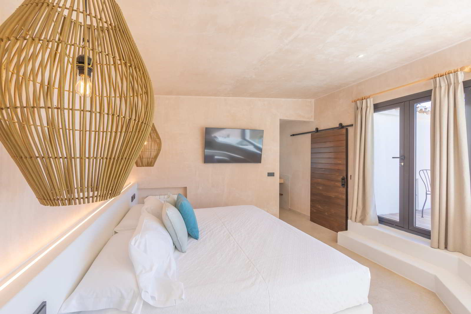 Dormitorio luminoso con cama doble, lámparas de fibras naturales, televisión y puerta corredera de madera en La Villa del Hotel Punta Sur.